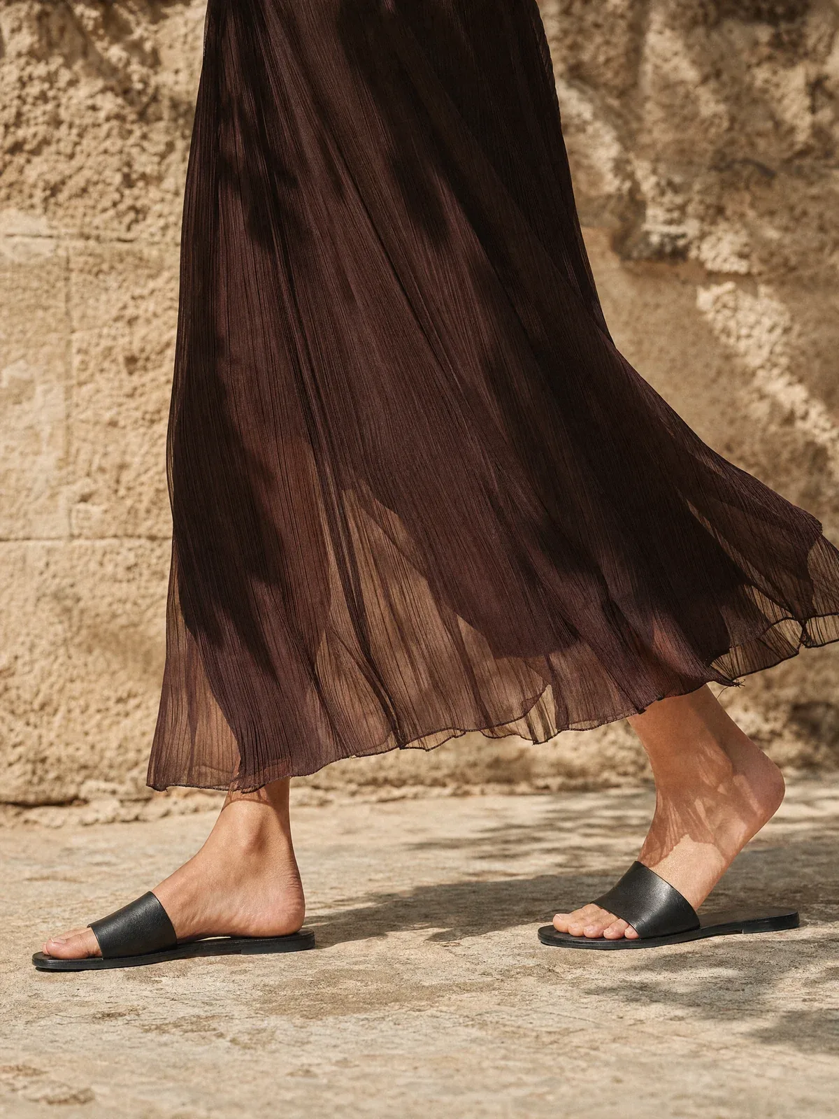 Modelo caminando con sandalias negras junto a una pared de piedra